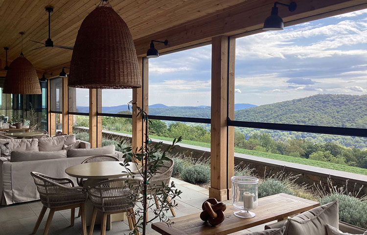 Open-sided covered patio or winery with retractable screens, wooden ceiling, pendant rattan lights, neutral upholstered furniture, and a panoramic view of a forested mountain valley